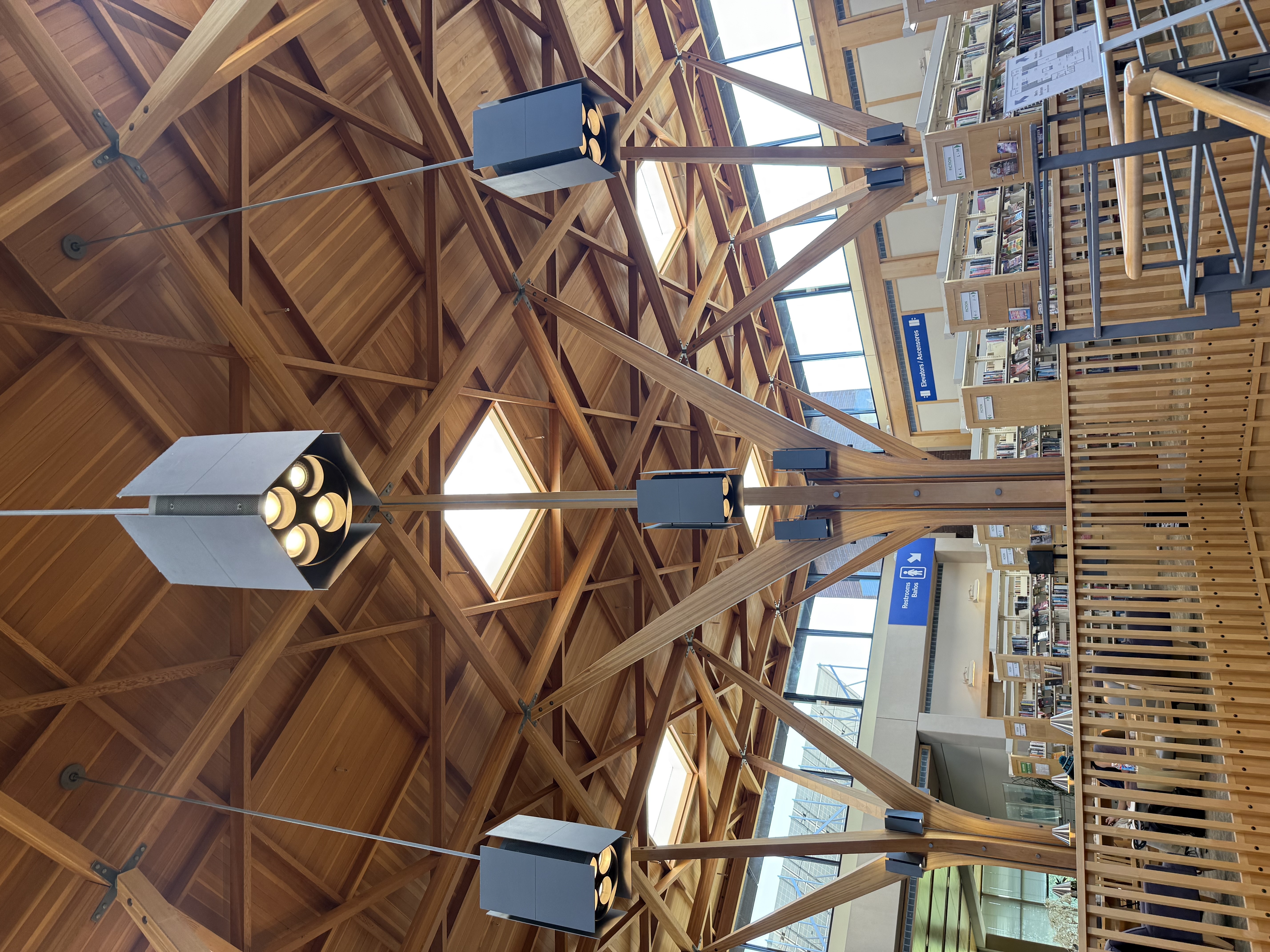 Looking up at the Beaverton City Library's diamond-grid glulam lattice ceiling, with pendant light fixtures and floor-to-ceiling glazing at the edges