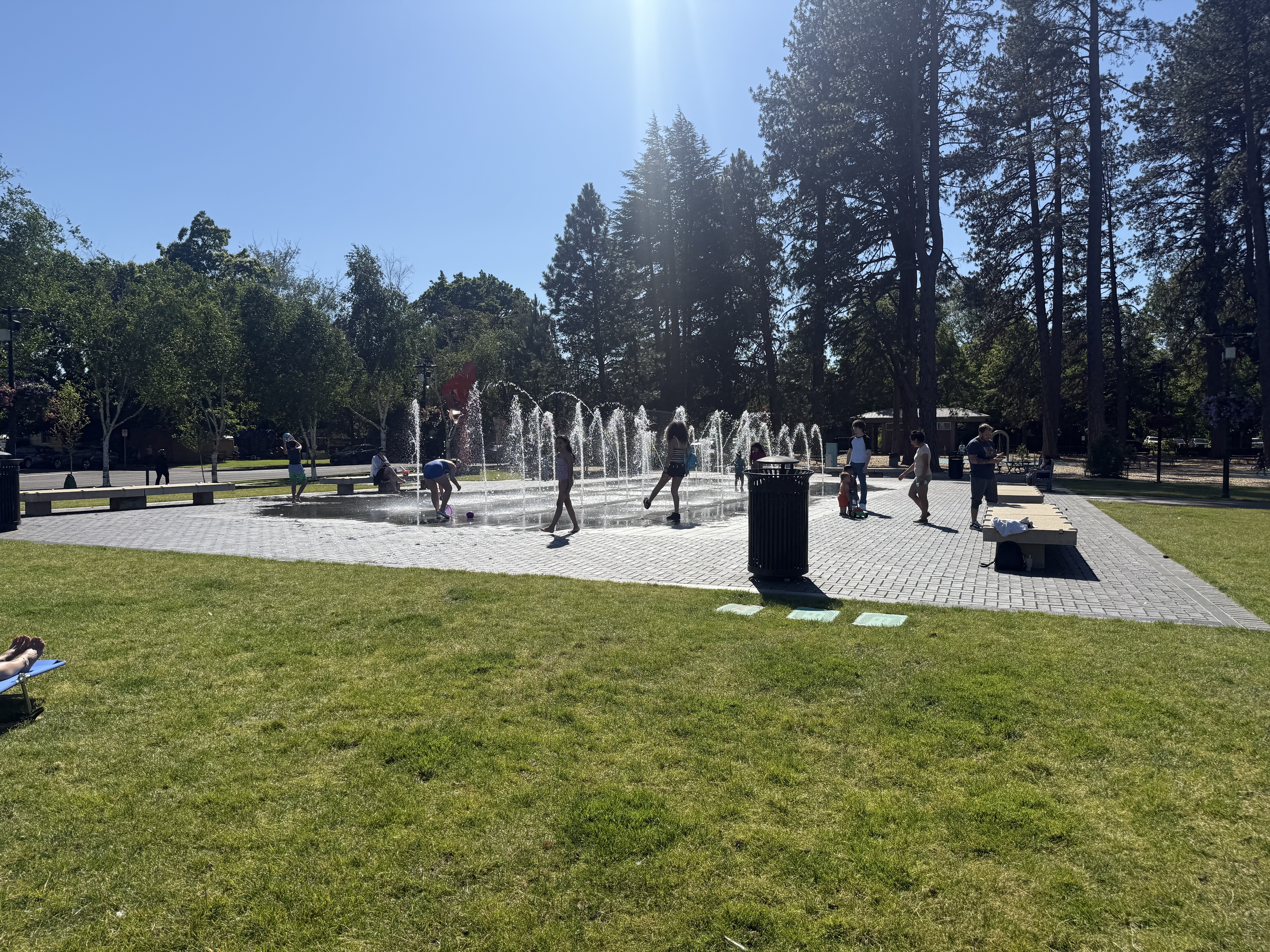 The Beaverton Civic Center splash pad — ground-level water jets on a paver surface, kids playing, tall conifers behind, benches and trash can visible