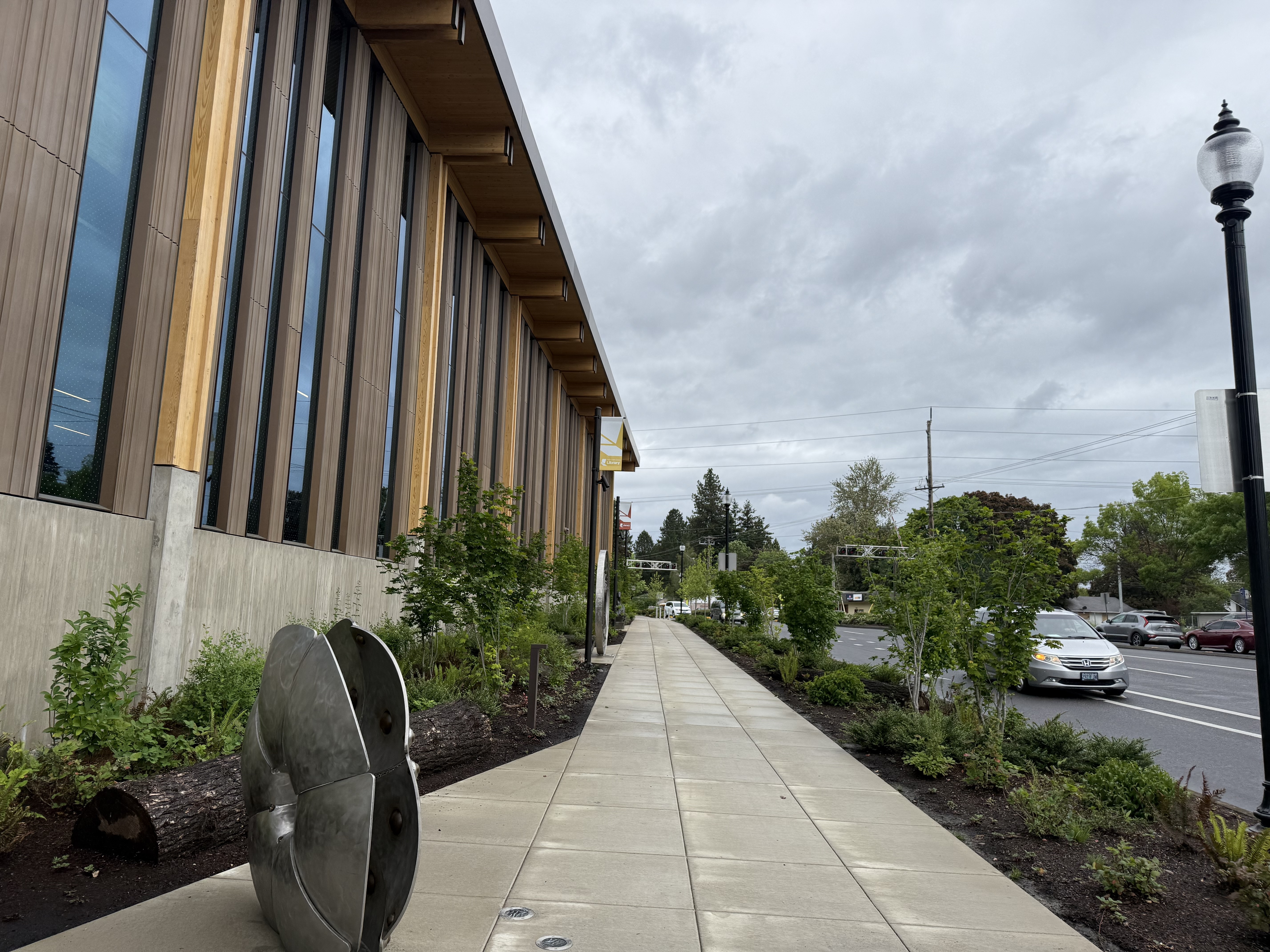 Full street-level view of the library exterior showing the timber building, new sidewalk plantings, and a metal disc sculpture in the foreground